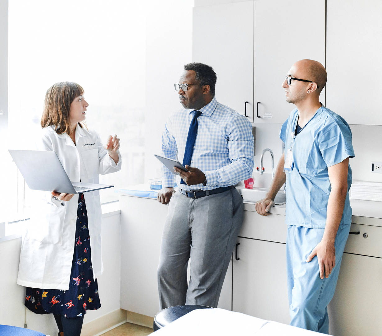 Three medical professionals (a light-skinned woman standing holding a laptop, a dark-skinned man leaning on the counter holding an electronic tablet, and a light-skinned man leaning on the counter next to the dark-skinned man) having a conversation in a doctor’s office.