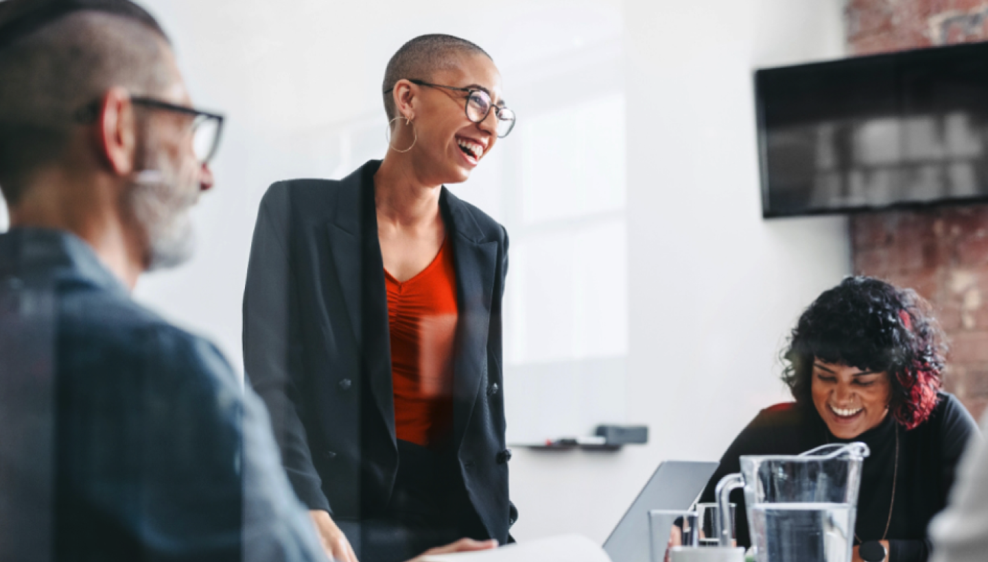 Three employees share a laugh during a meeting. Two are seated and one is standing. 