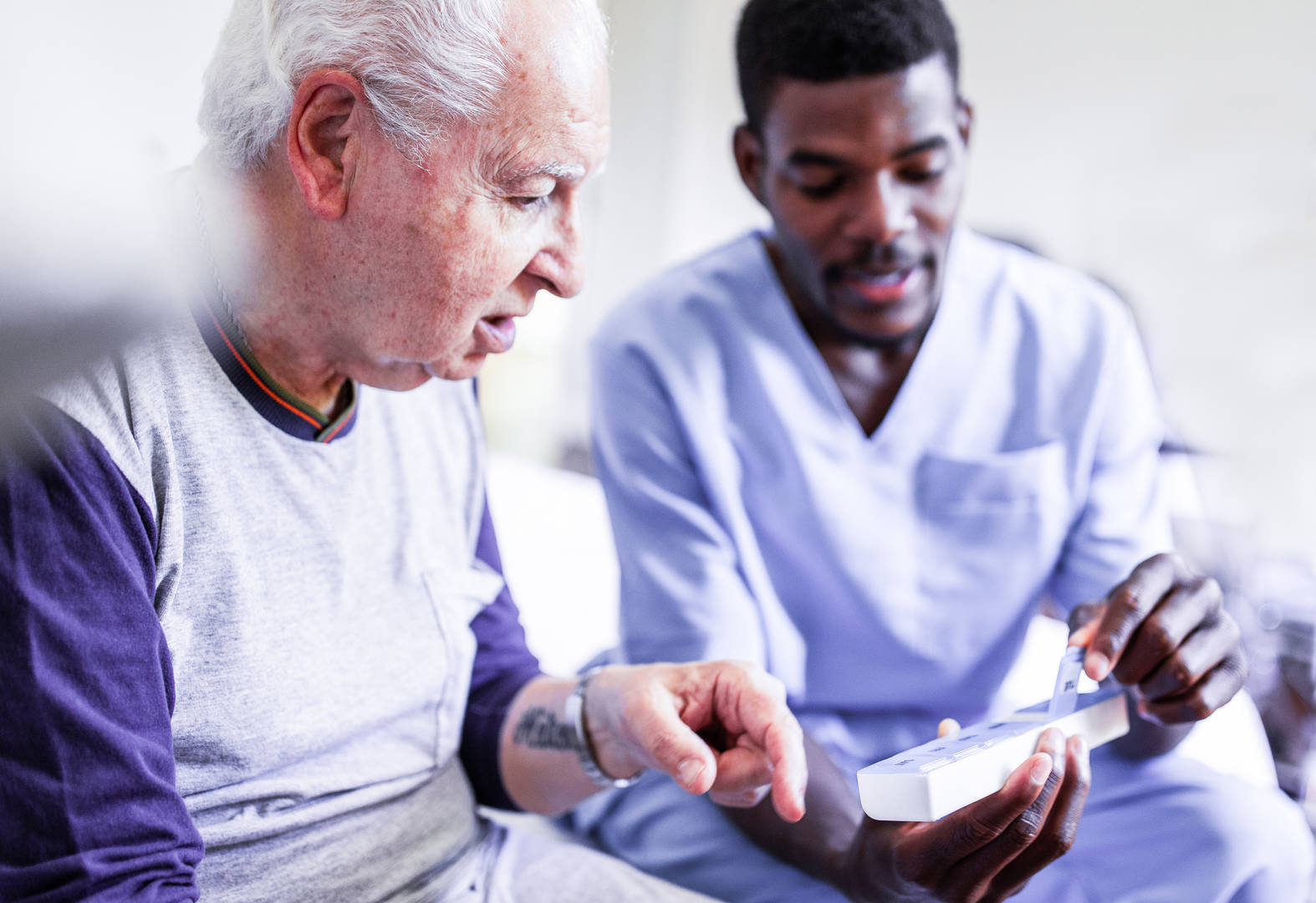 A medical professional wearing blue scrubs sits beside a patient in a long-sleeved shirt, explaining the medication dosage.