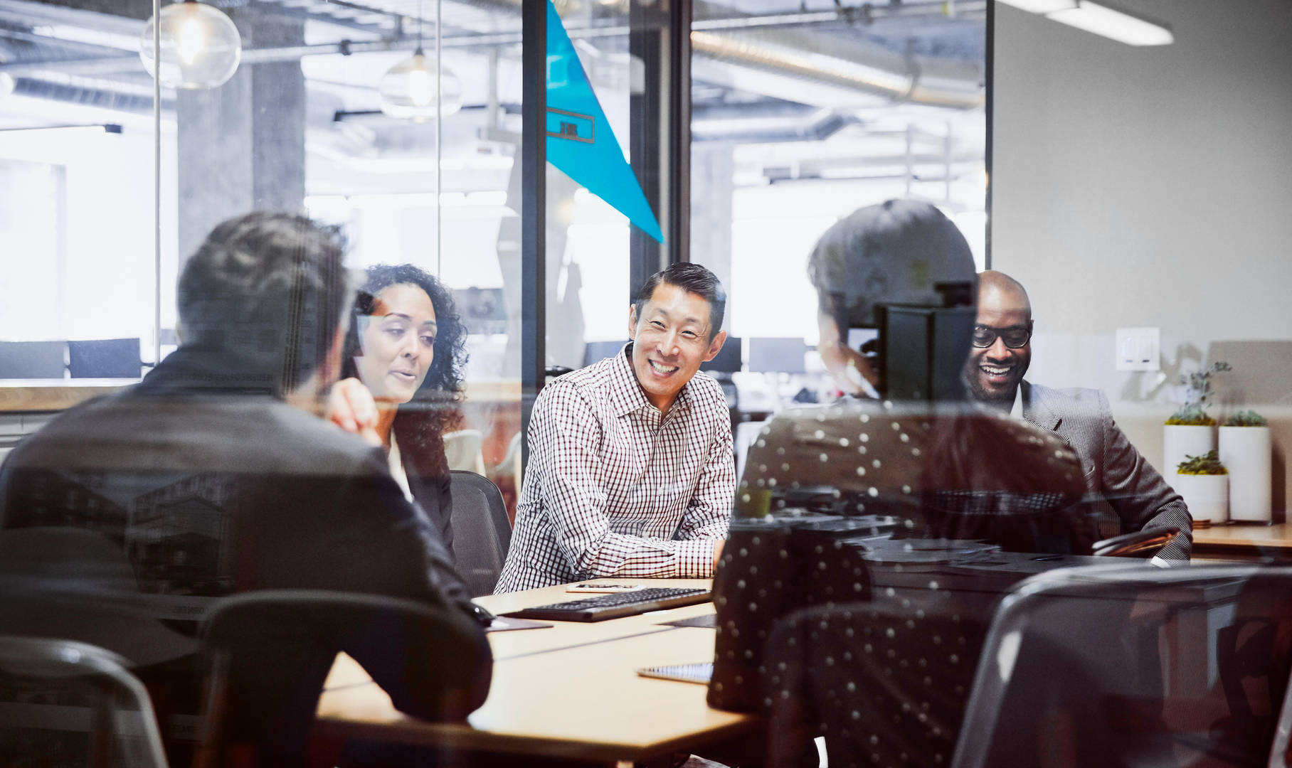 Four coworkers sit in an office with large glass windows, wearing business casual clothing and smiling in a meeting.