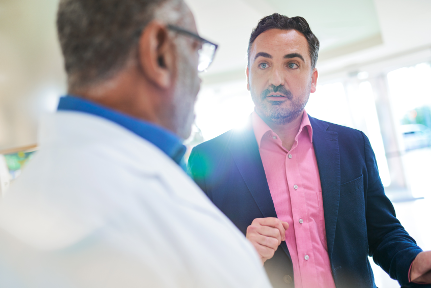 A salesperson wearing a dark suit with a bright shirt talks to a pharmacist wearing a white coat at a local pharmacy.
