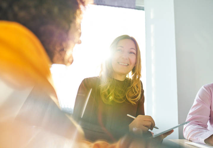 A light-skinned person with long brown hair holding a tablet and stylus smiles at a colleague during a meeting in a conference room.