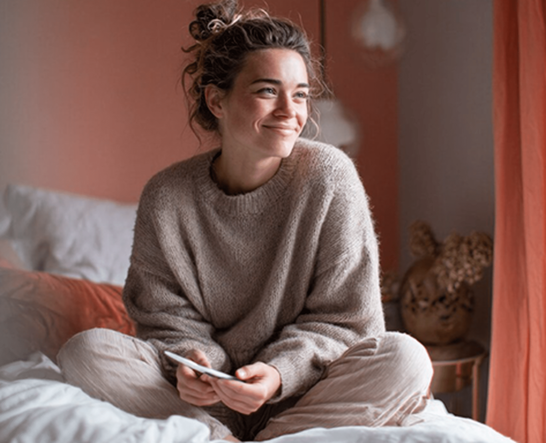 Smiling your woman sitting on a comfortable bed and holding a phone in her hands