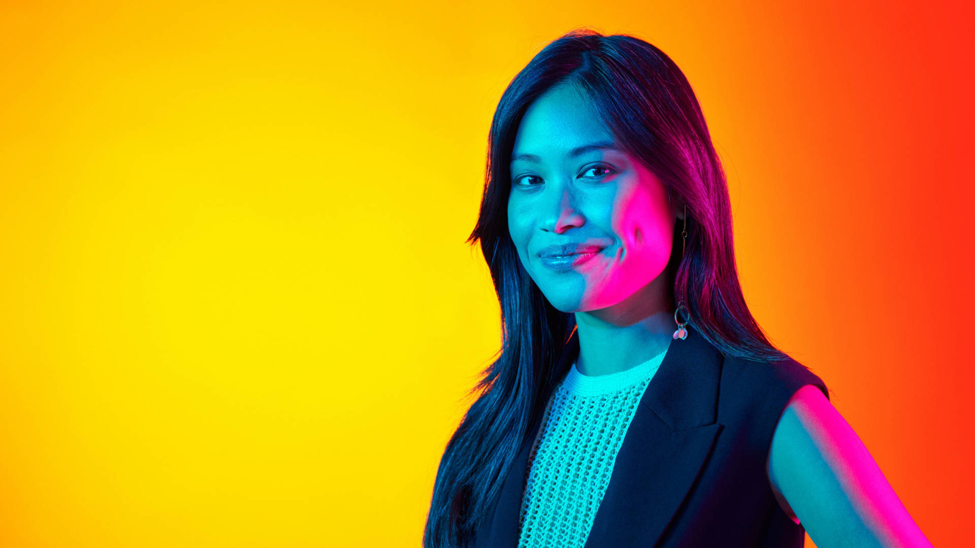 A woman is smiling at the camera against an orange and yellow background. Her face is lit with blue and magenta lights.