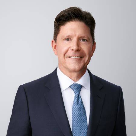 Photograph of Robert P. Mauch. Robert, a light-skinned person with black hair and green eyes, is smiling at the camera. He wears a black suit overtop a white shirt and blue necktie. He is in front of a light grey background with no surroundings.