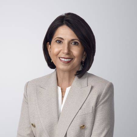Photograph of Silvana Battaglia. Silvana, a light-skinned person with brown hair and brown eyes, is wearing earrings and smiling at the camera. She wears a gray blazer jacket over a white shirt, and a necklace. She is in front of a light gray background with no surroundings.