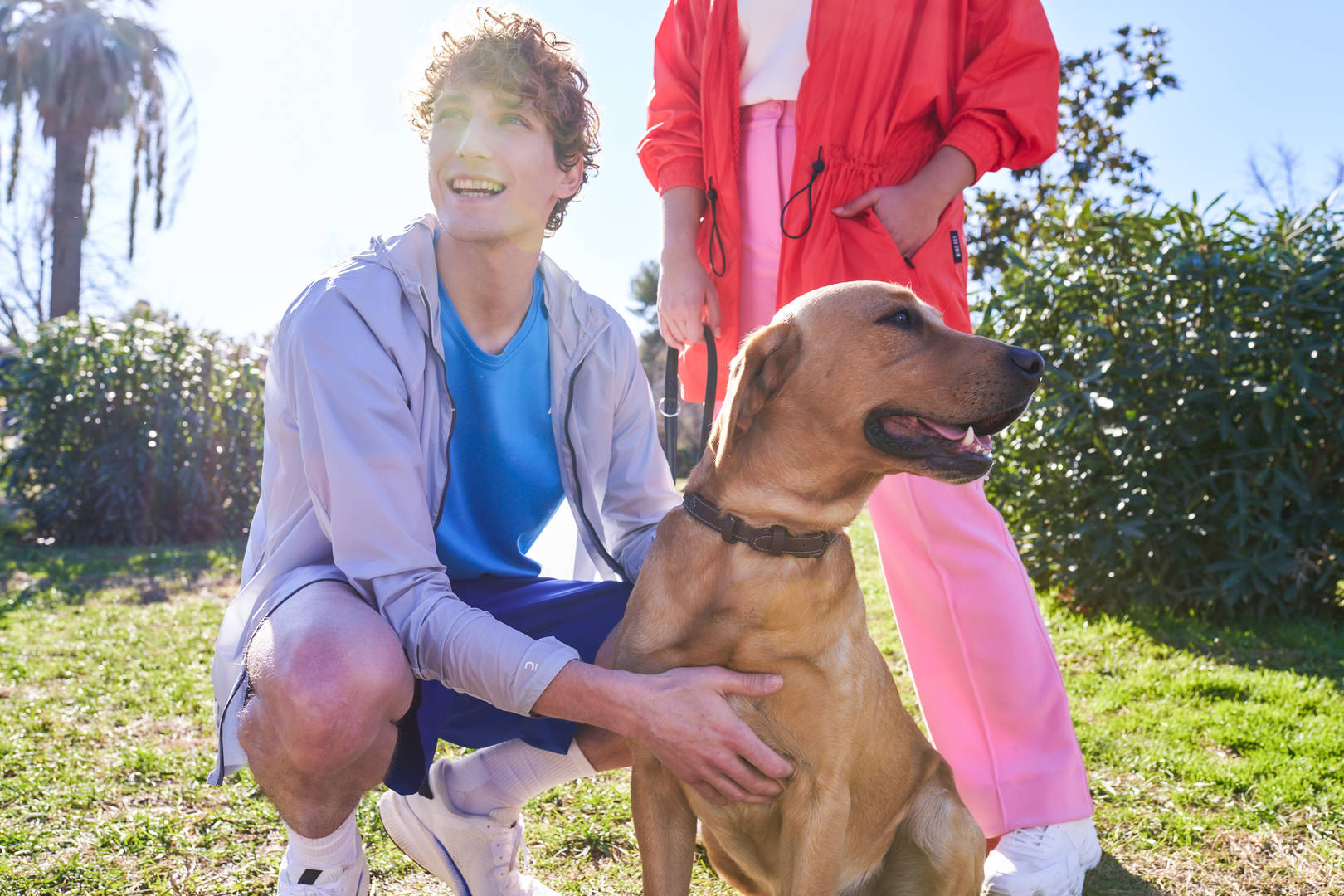 Un jeune homme s’accroupit pour caresser un chien tandis qu’une femme se tient en arrière-plan, tenant la laisse du chien.