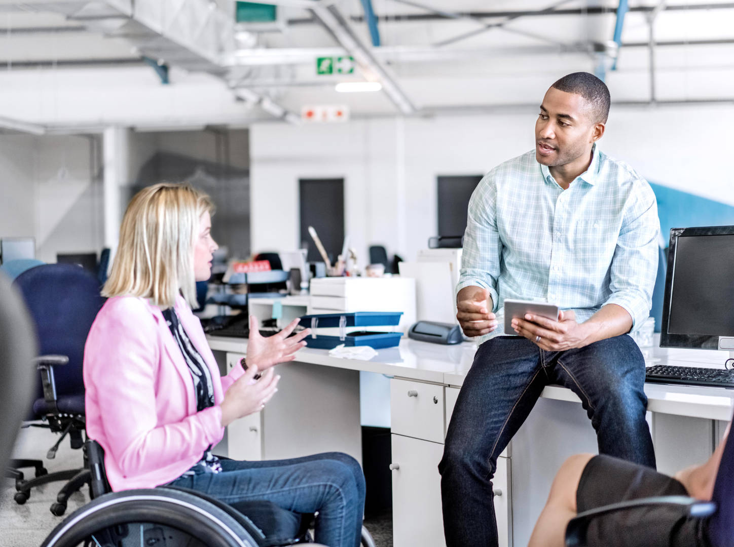 Two co-workers (a light-skinned person with shoulder-length hair sitting in a wheelchair and a dark-skinned person with short hair sitting on the edge of a desk) wearing business casual clothing talk in their office.