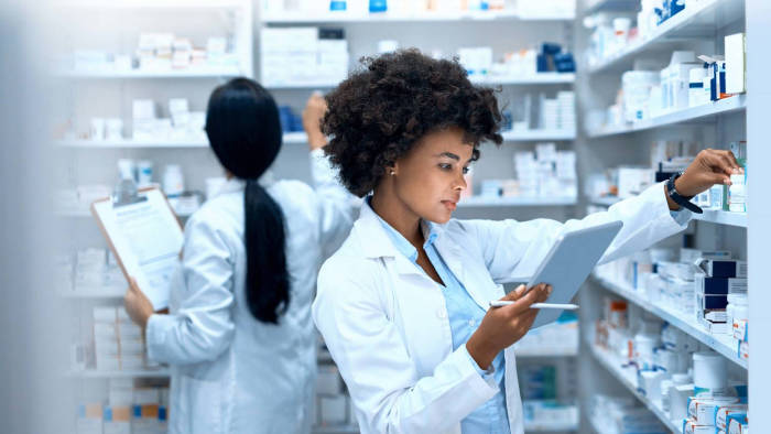 Two pharmacists pulling medications from a pharmacy shelf. They both wear white lab coats. One has curly, dark hair and dark skin and holds a tablet device. The other has long, straight, dark hair and has their back turned to the camera while holding a clip board.