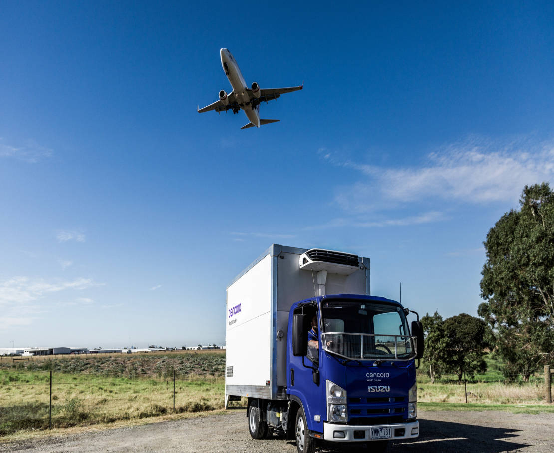 A blue and white box truck with the Cencora logo printed on it drives past a grassy field while an airplane flies overhead during a sunny day.