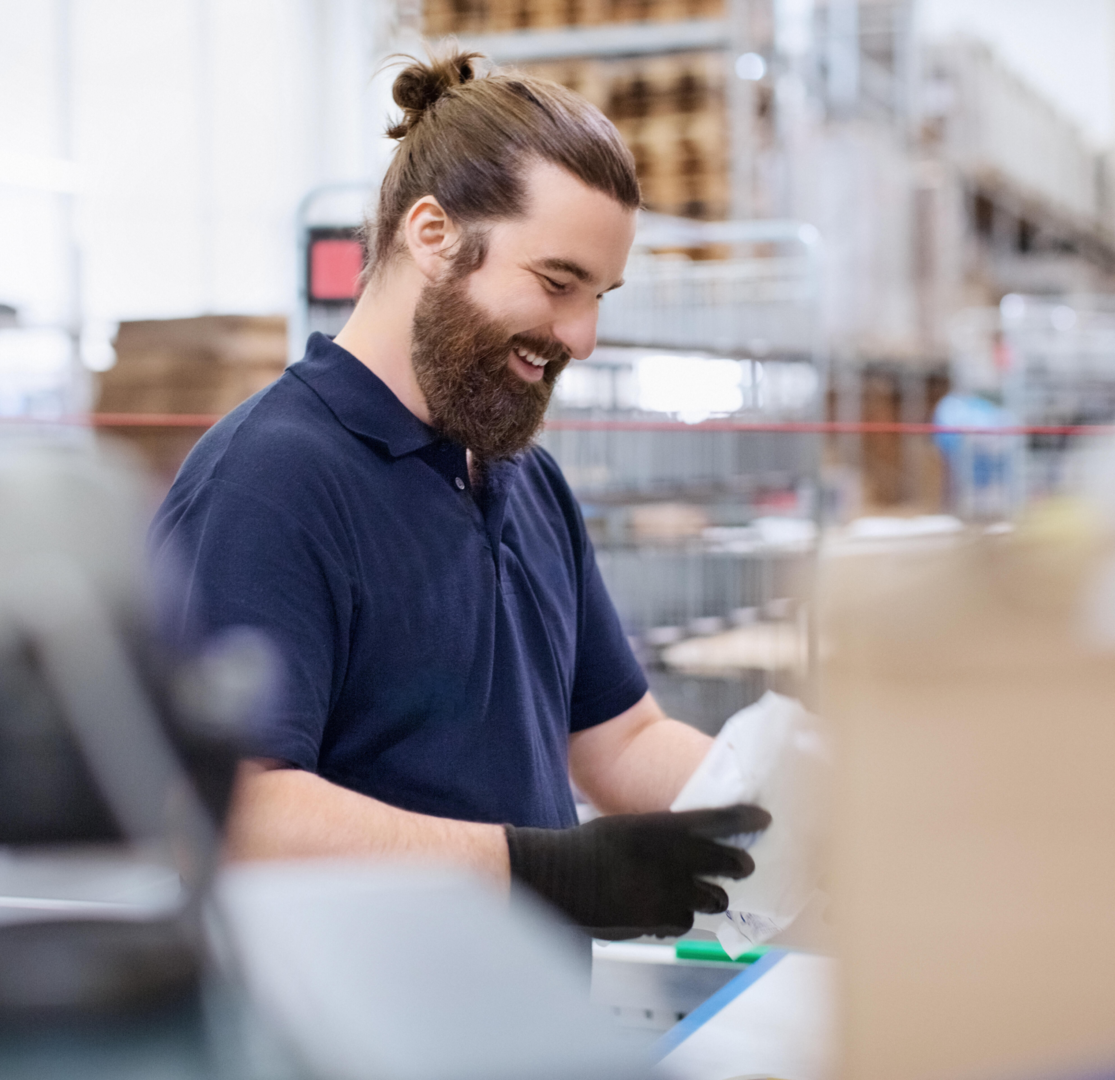 Un employé d’entrepôt charge des colis dans des caisses dans un centre de distribution. Ils ont de longs cheveux bruns attachés en chignon, une barbe, et ils portent un polo bleu marine et des gants noirs.