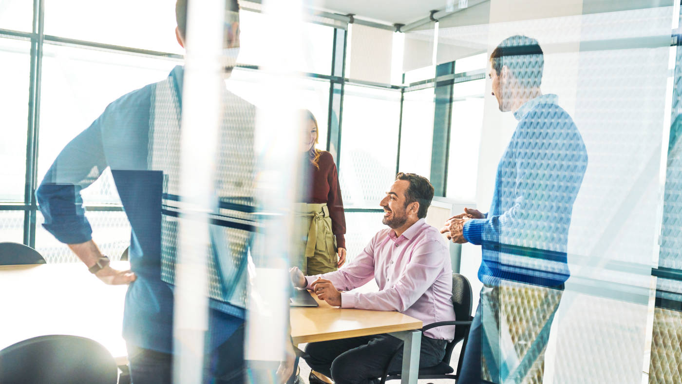 Vier Menschen lächeln und lachen an einem Tisch in einem hellen Konferenzraum. Drei von ihnen stehen am Tisch, einer sitzt mit einem Laptop vor sich auf dem Tisch.