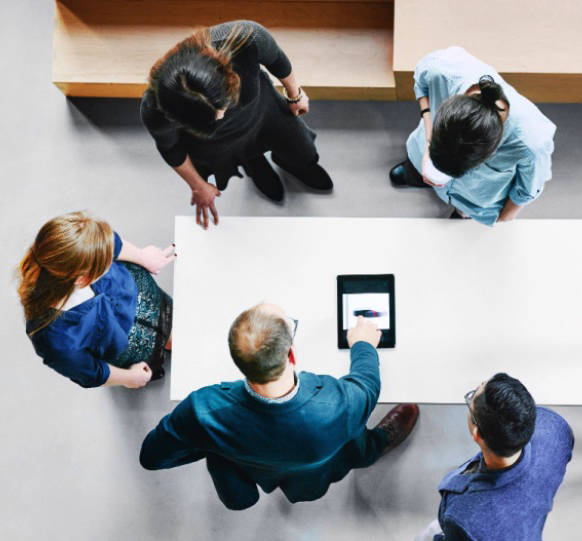 An overhead view of five employees looking at a tablet while standing around a table.
