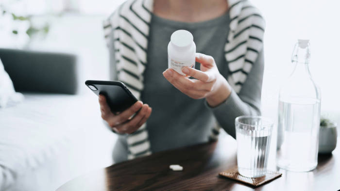 A person holding a white prescription medication bottle in one hand and a cell phone in another. They sit at a table with a glass of water in front of them while wearing a gray long-sleeve shirt and a black and white striped sweater.