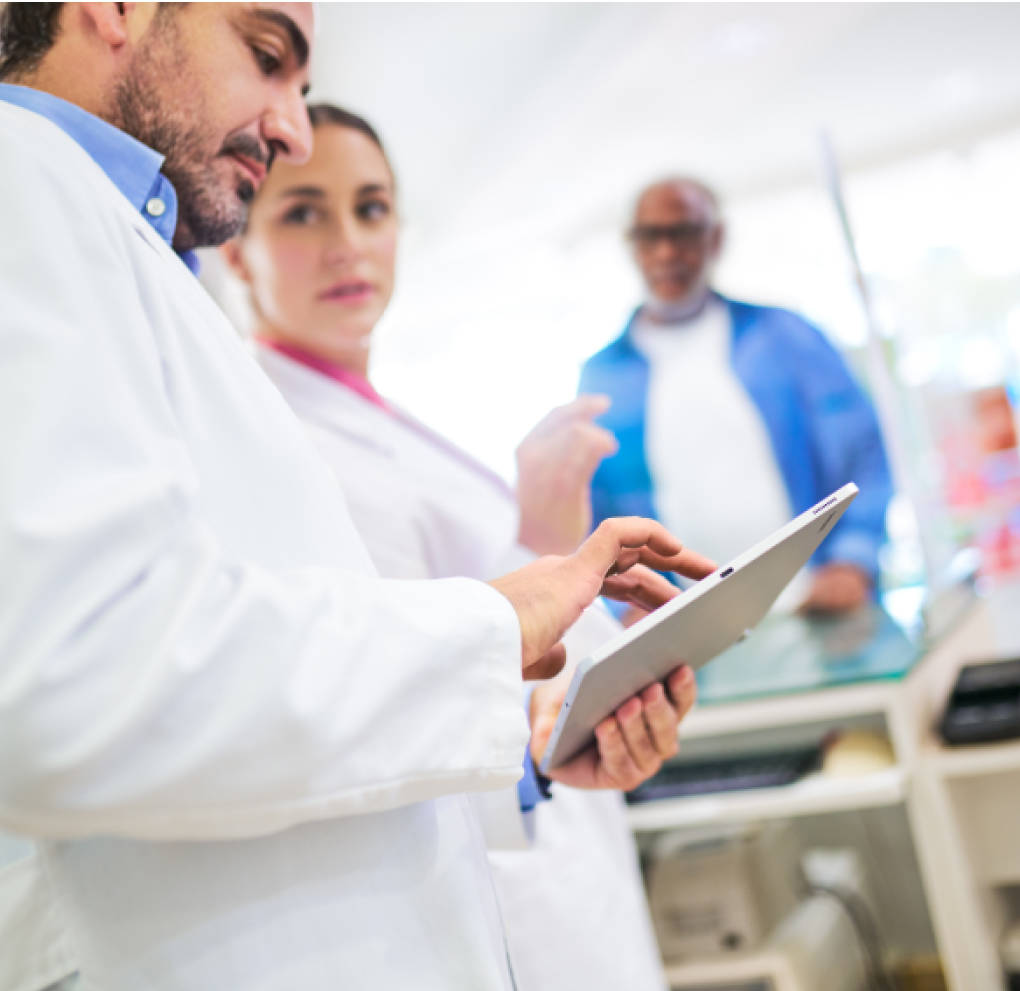 A pharmacist holding a tablet device consulting with another pharmacist while a patient waits behind the pharmacy counter.