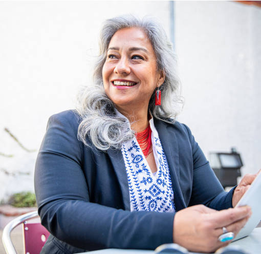 A person sitting at a table looks over their right shoulder and smiles past the camera. They have long gray hair and wear red earrings, a blue sweater, and a white and blue blouse. They hold and operate a tablet device.