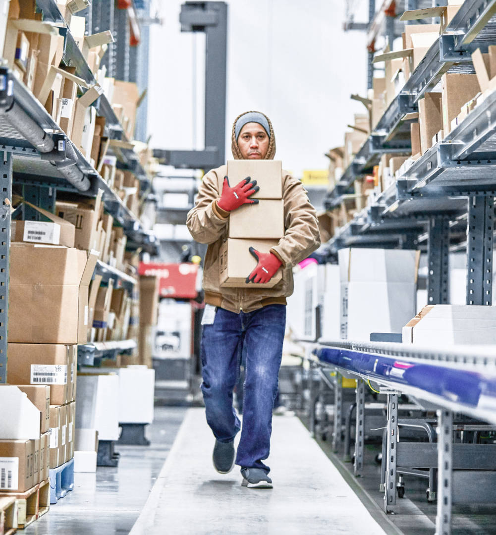 A warehouse worker wearing red gloves carrying three medium-sized boxes down an aisle in one of our distribution centers.