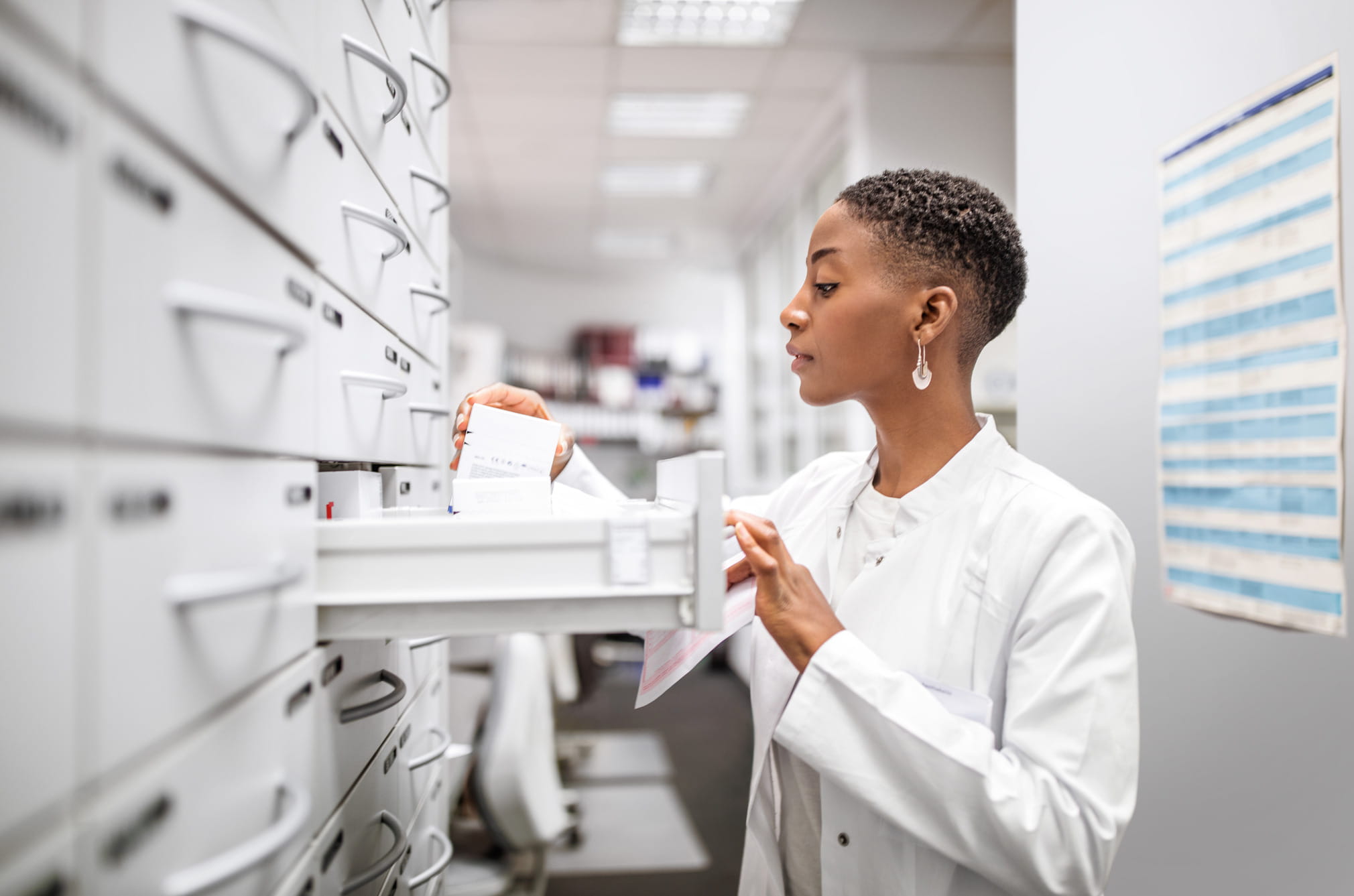 A pharmacist checks a prescription while retrieving a medication from pharmacy storage drawers.
