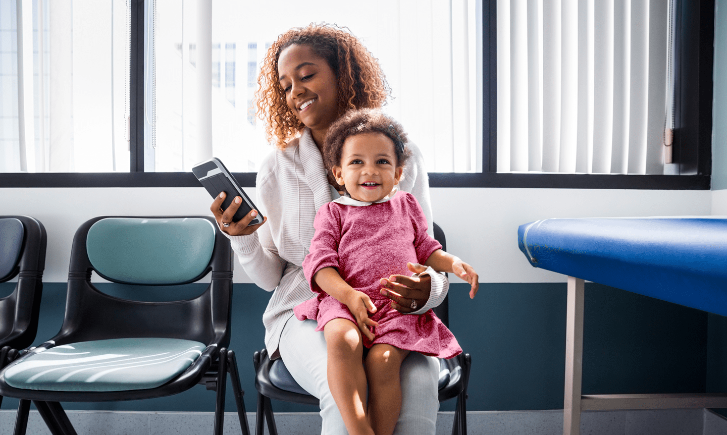 A mom checking her phone with her daughter smiling and sitting on her lap in an exam room.