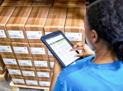 A woman wearing a blue t-shirt is reviewing inventory data on a small tablet device while standing in front of a pallet of boxes in a distribution center.