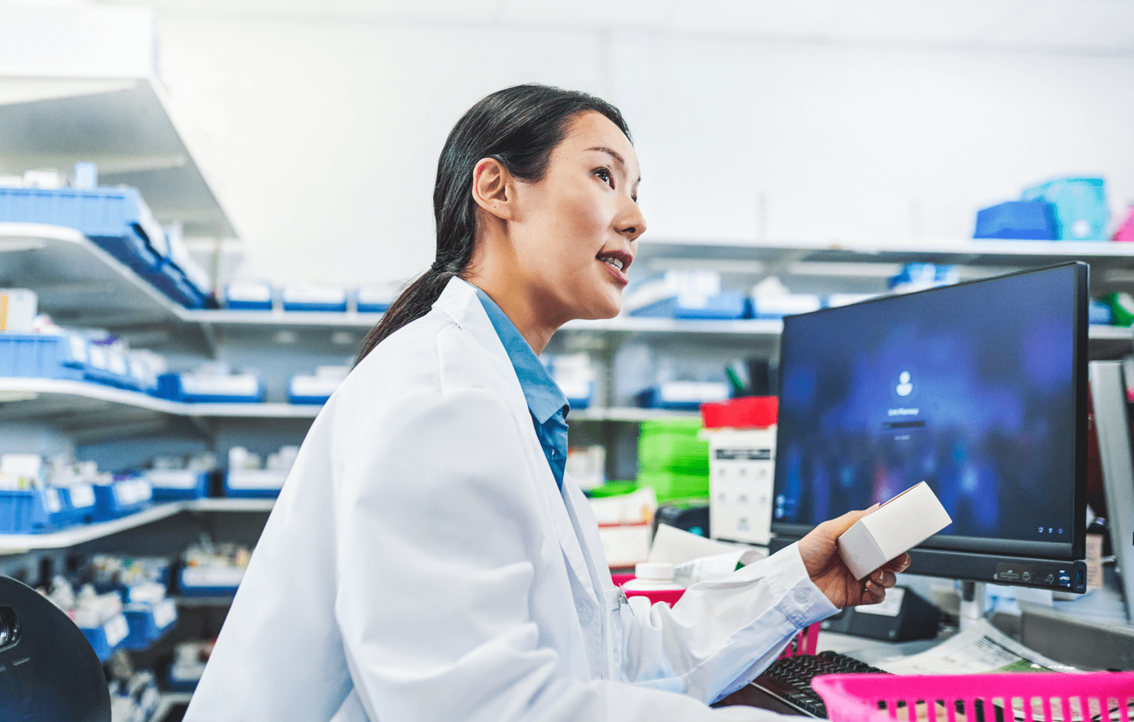 A female pharmacist wearing a white lab coat is standing near a computer monitor behind a pharmacy counter while holding a box of medicine. Behind her, there are several shelves filled with pharmacy inventory.