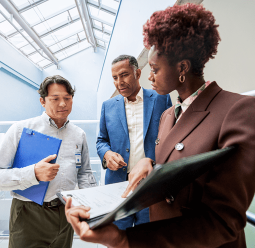 Two men and a woman are having a discussion in a brightly lit hallway. They are all wearing business professional attire. The woman is holding a portfolio folder and pointing to the documents inside.