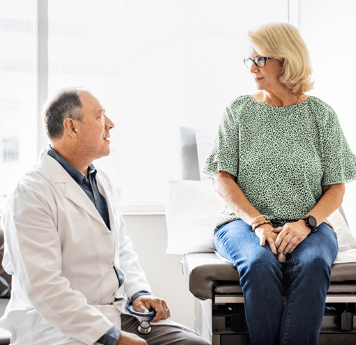 A senior male doctor is consulting a senior female patient in an exam room. The doctor is sitting on a stool on the left, and the woman is sitting on an exam table on the right.