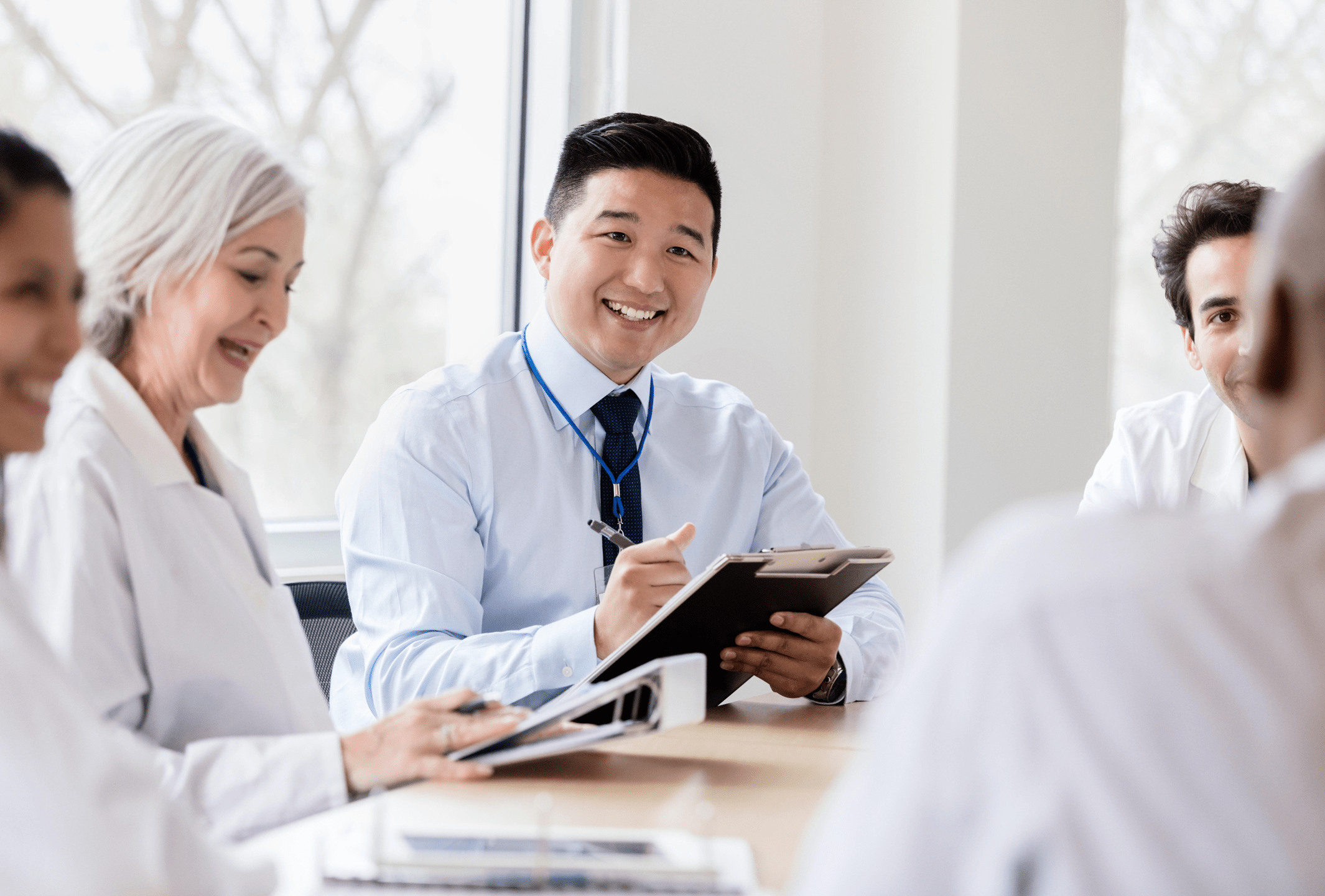 A multicultural mixture of smiling women and men sit at a conference table. There are business-related items in their hands and on the table. 