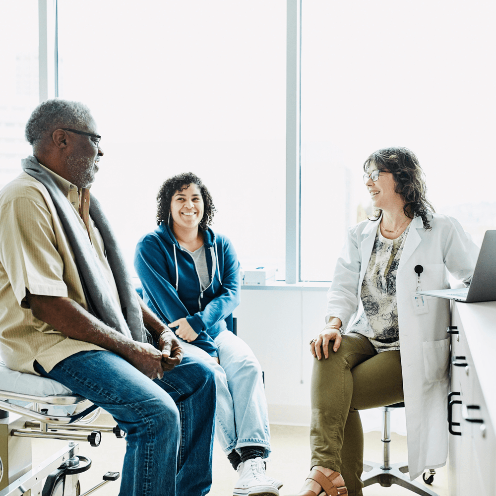 A light-skinned woman in a lab coat consults with a seated dark-skinned man wearing blue jeans and a yellow short-sleeved shirt and a seated light-skinned woman wearing blue jeans and a blue shirt in a consultation room.