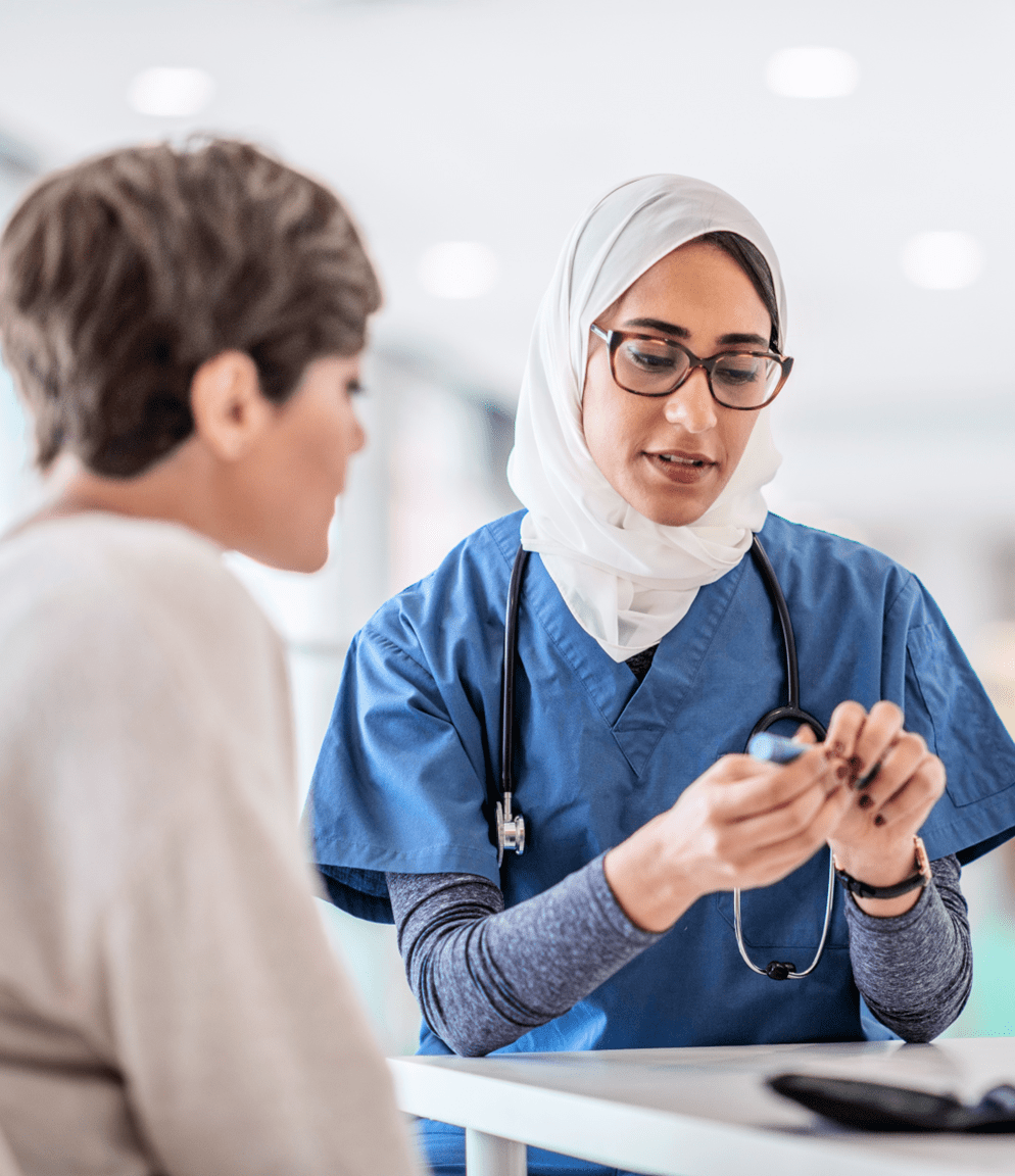 A medium-light-skinned woman wearing dark eyeglasses and a white hijab shows a light-skinned woman wearing a light brown sweater a vial of medicine.