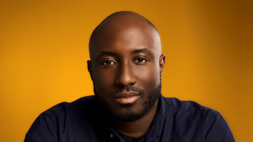 A studio portrait of a man looking forward with a slight smile. He is wearing a black shirt in front of a gold background.