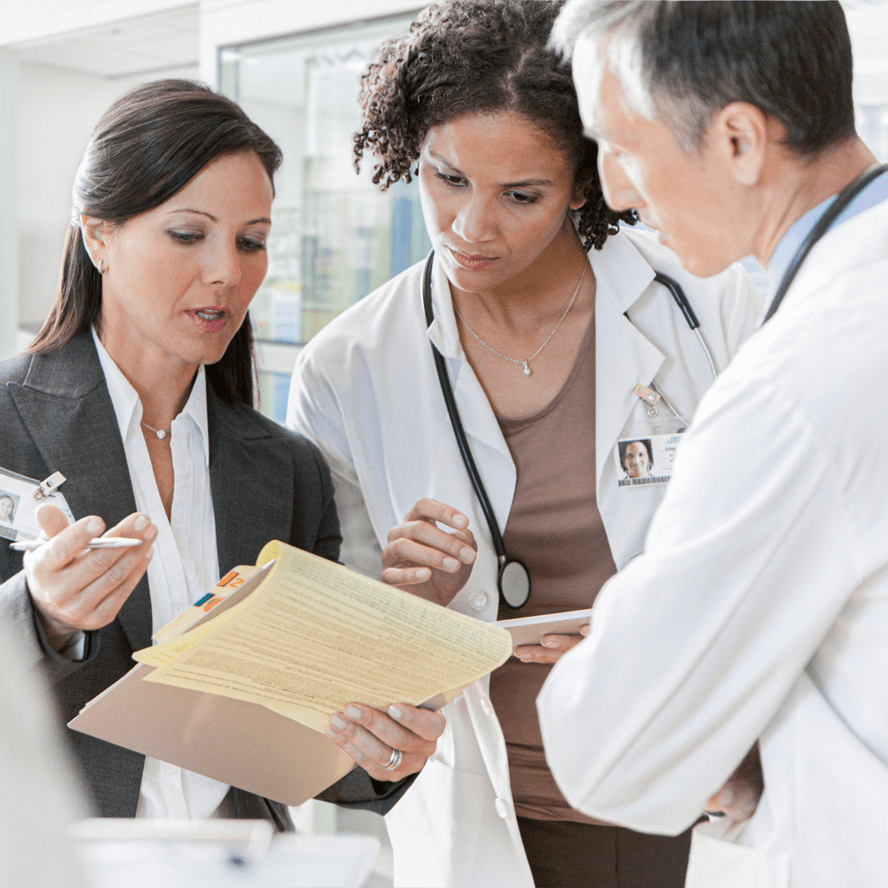 A woman in business attire is holding a yellow notepad and talking to a male doctor and a female doctor who are both wearing white lab coats.