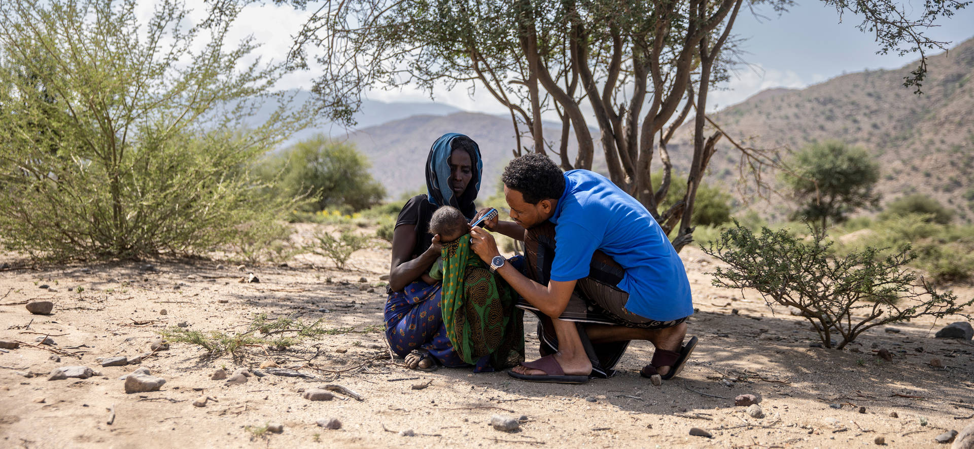 A casually dressed man in sandals is conducting a diagnostic test on a baby, which is being held by its mother. They are crouching on the ground in a rural setting.