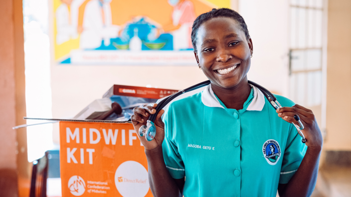 A medical professional wearing teal scrubs and a stethoscope is standing in front of an orange box labeled “midwife kit” that is filled with medical supplies.