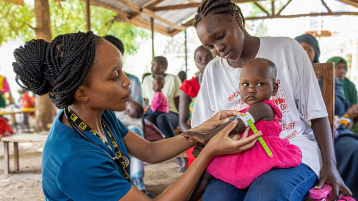 A medical professional wearing blue scrubs and a stethoscope is evaluating a toddler’s arm with a tape measure. The child is wearing a pink dress and sitting on their parent's lap.