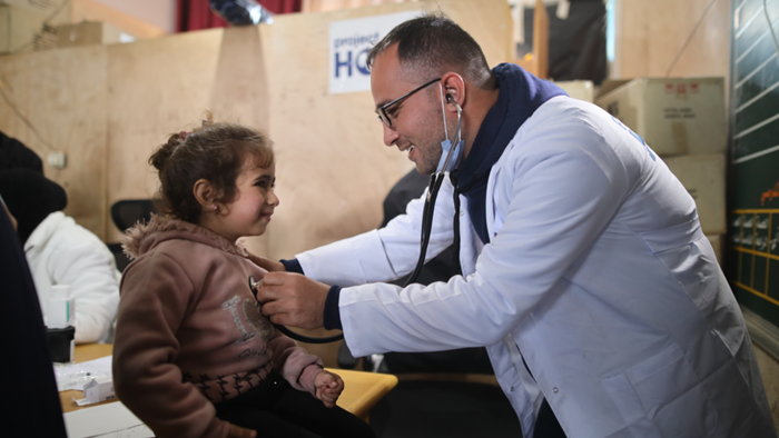 A medical professional is using a stethoscope to check the heartbeat of a child who is sitting on a table. The medical professional is wearing a white coat on top of a dark blue hooded sweatshirt, and the child is wearing a tan hooded sweatshirt.