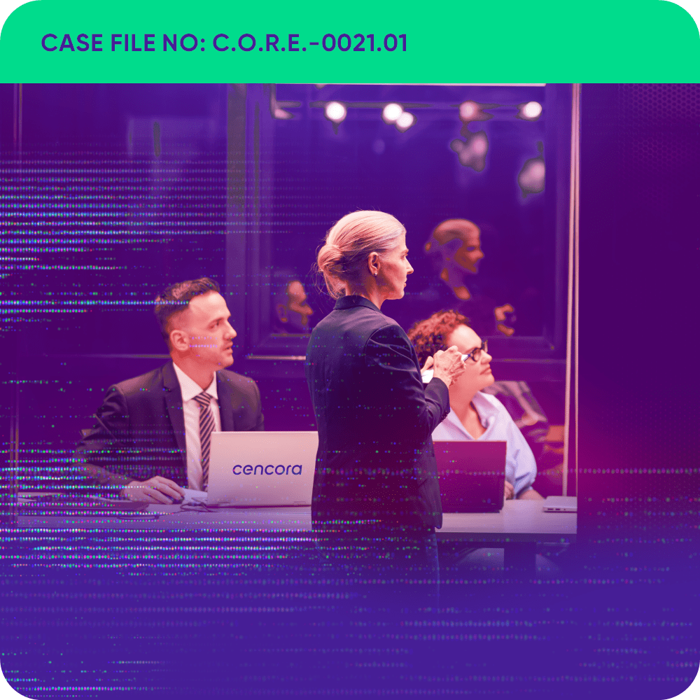 A man and a woman wearing business professional attire are sitting at a meeting room table with their laptops open in front of them. A woman wearing a suit is standing on the other side of the table. All three are looking right and watching a presentation.