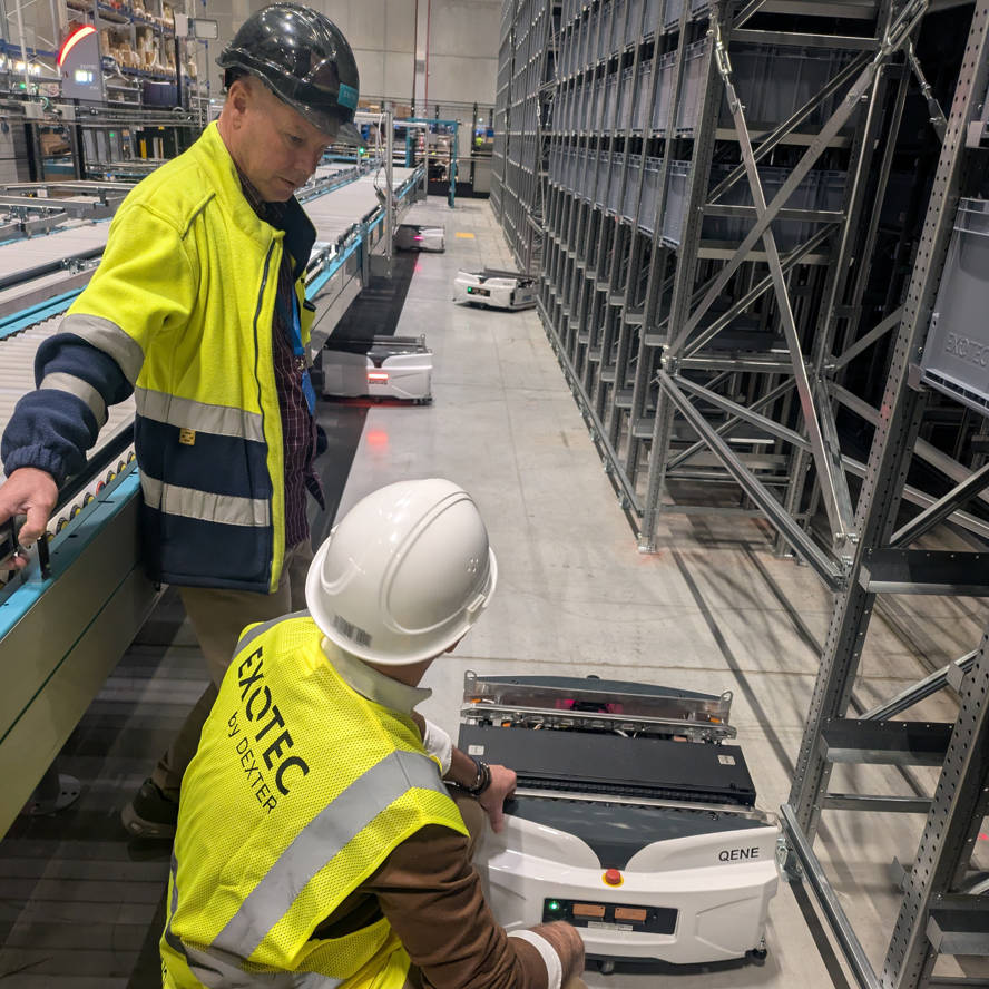 Two men wearing hardhats and yellow safety vests examine an autonomous warehouse robot. One man is standing, the other is crouching next to the robot.