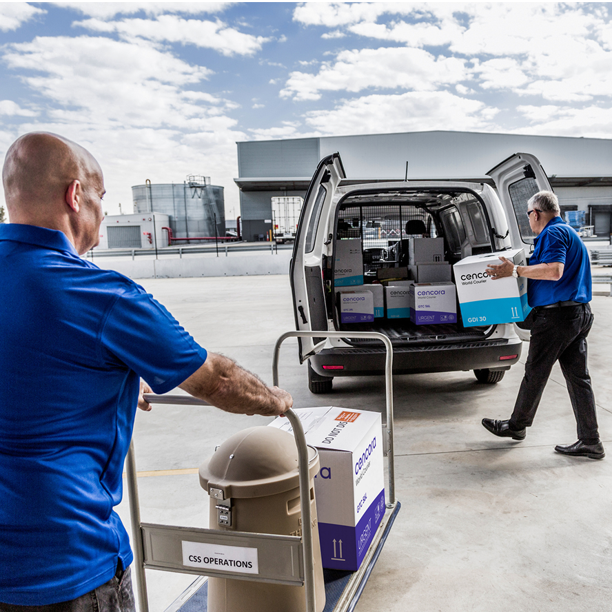 Two men load containers into a Cencora delivery van. One pushes a fully-loaded flatbed cart, while the other totes a large box manually.