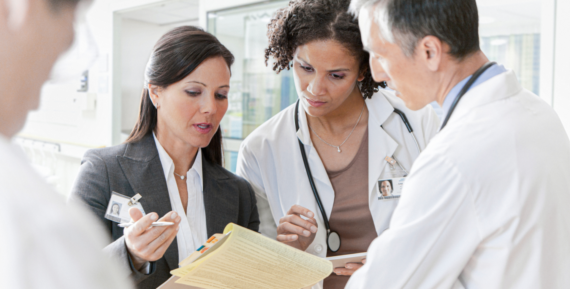 A business professional and three medical professionals in white lab coats huddle together reviewing documents from a manila folder in a hospital setting.