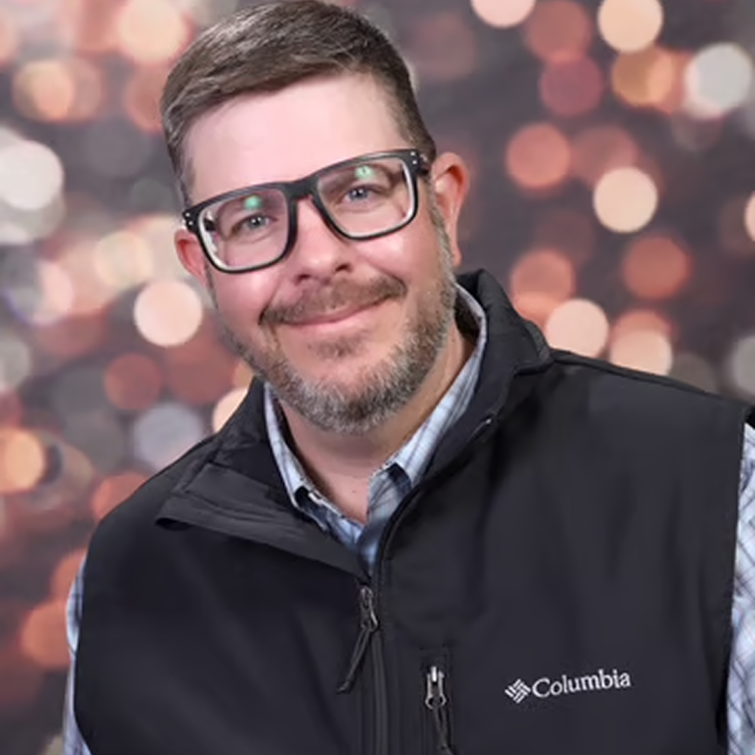 A headshot of a Supervisor at Cencora, a middle-aged man with short brown hair, black prescription glasses, and a slight smile, dressed black vest over a button down, conveying leadership and professionalism.