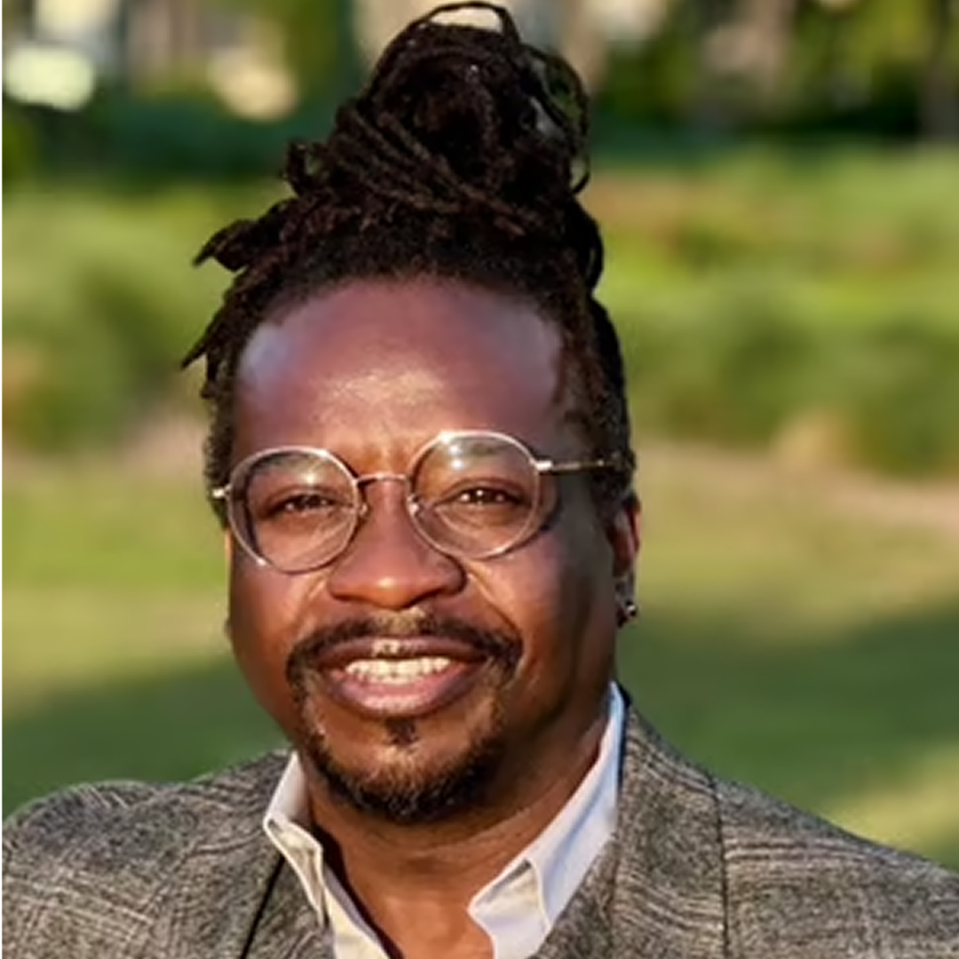 A headshot of a Project Manager at Cencora, a middle aged man with long hair tied on top, round silver prescription glasses, and a slight smile, dressed in a gray suit conveying leadership and professionalism.