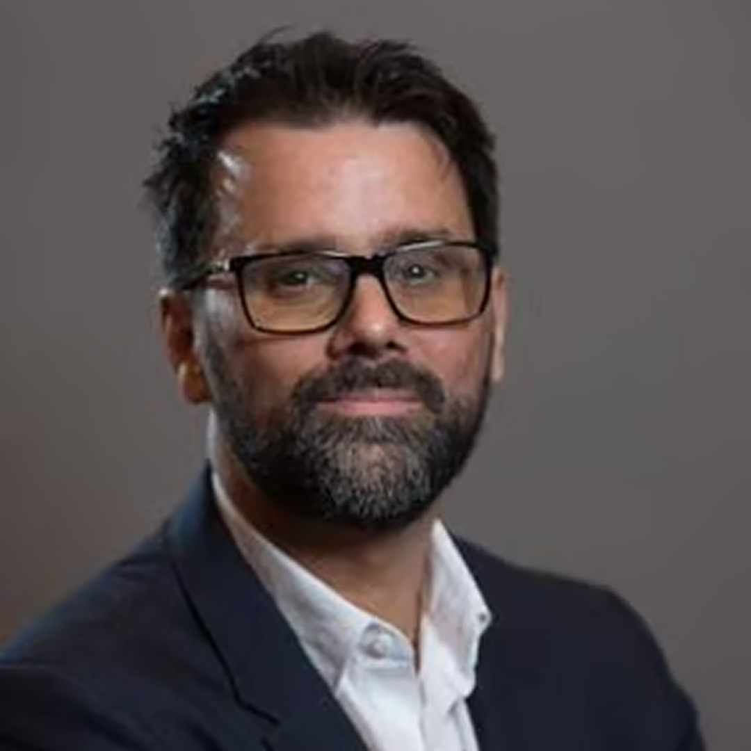 A headshot of a Director at Cencora, a middle aged man with medium length hair, rectangular black prescription glasses, and a slight smile, dressed in a blue suit conveying leadership and professionalism.