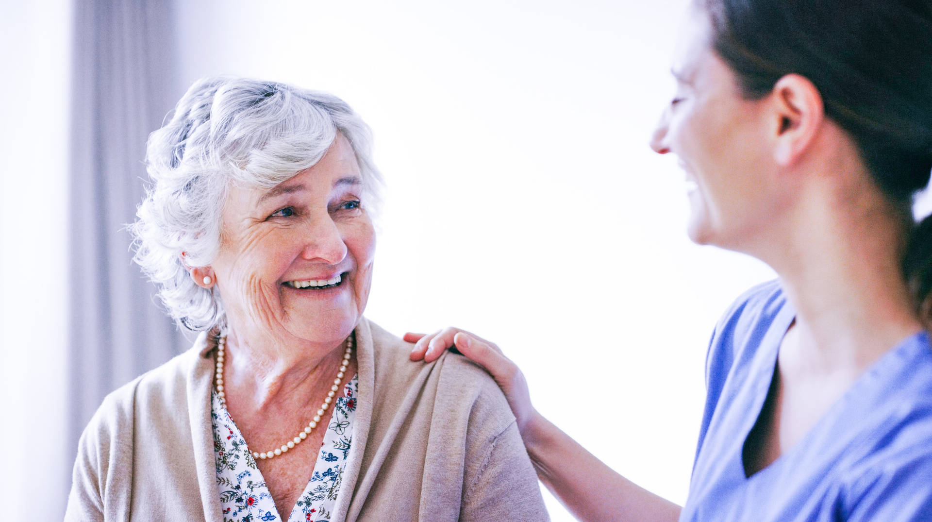 Care professional standing beside an older adult indoors, with a hand resting on the older adult’s shoulder during a conversation.