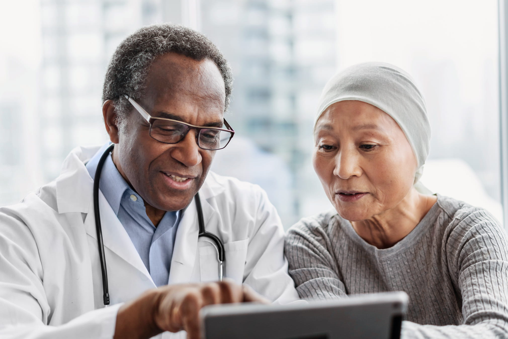 Healthcare professional in a white coat with a stethoscope shows information on a tablet to a patient wearing a headscarf, seated together in a clinical setting.