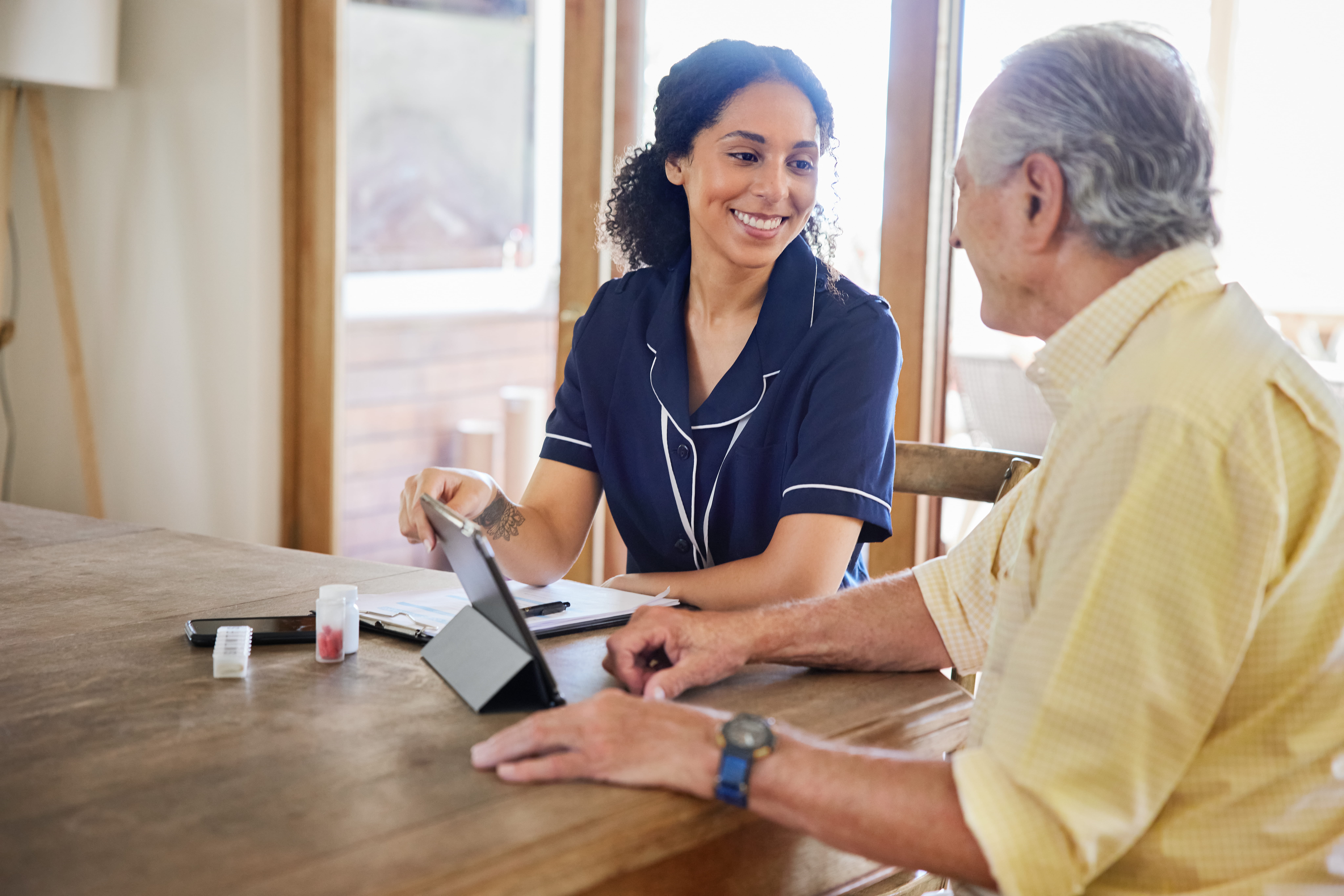 A female homecare nurse is visiting a patient and they are discussing treatment on the dining room table with medications and a tablet. 