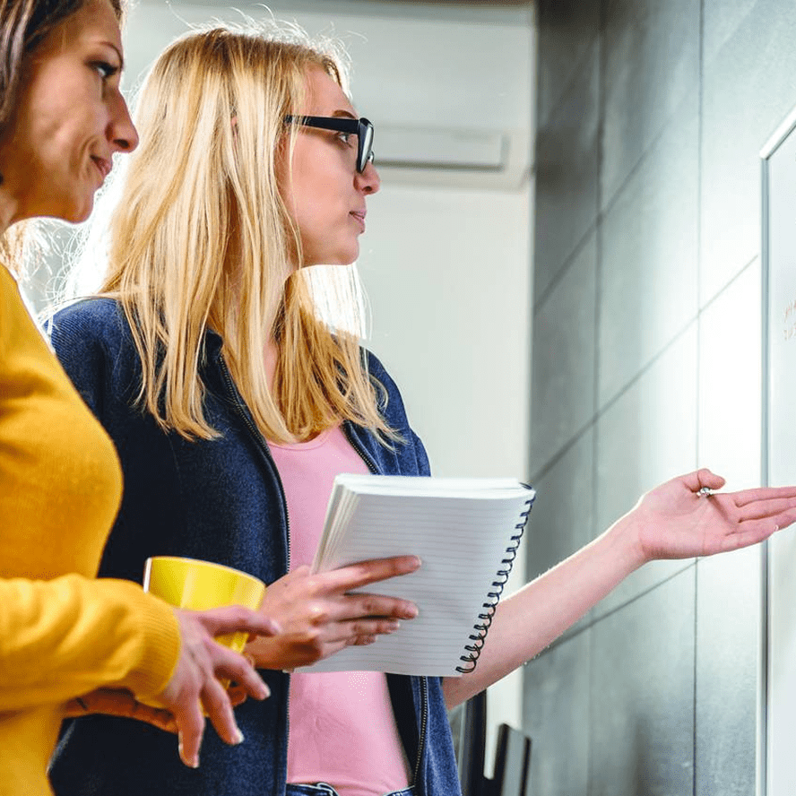 two women looking at a board, holding a notebook and a mug