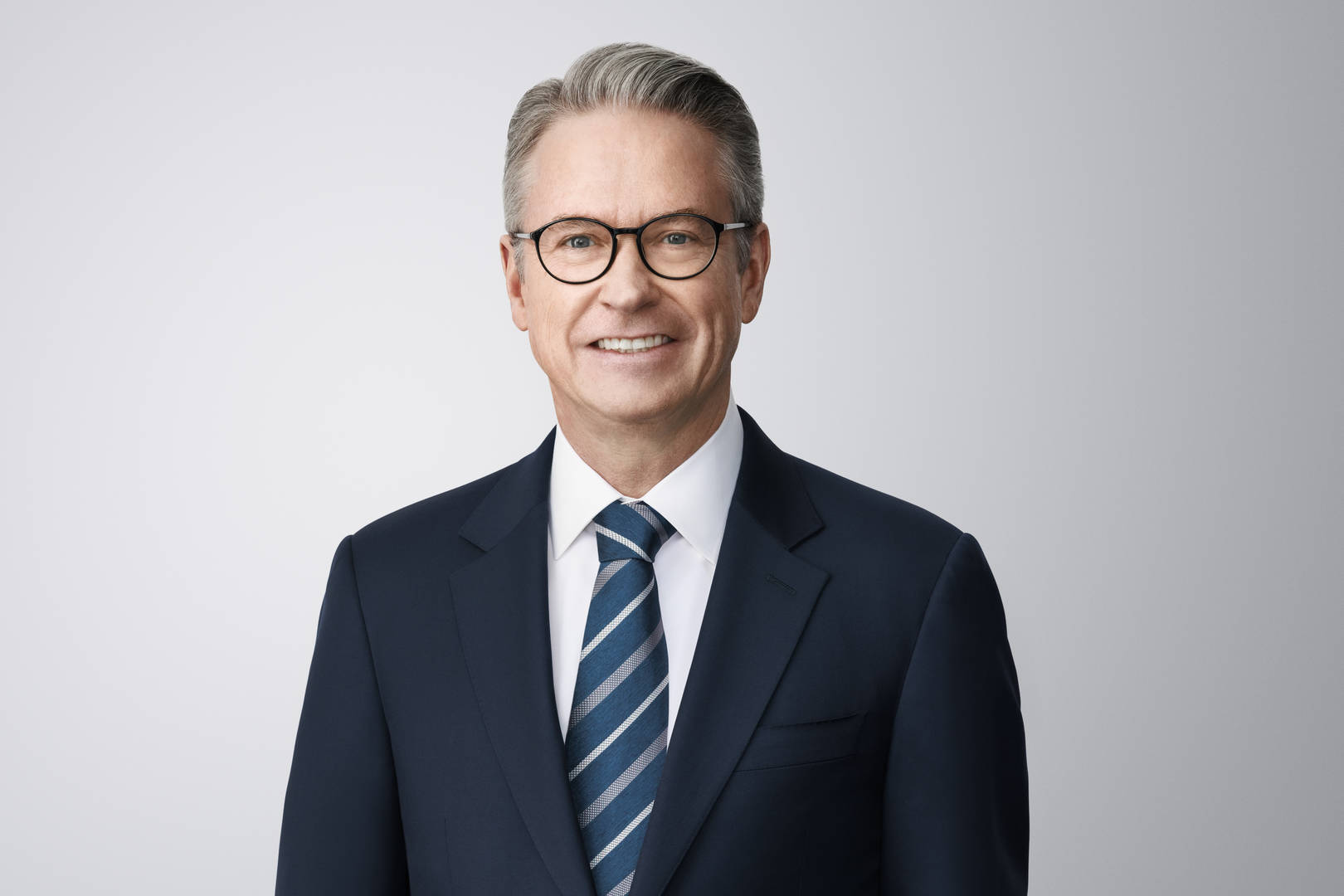 Foto von James F. Cleary. James, eine hellhäutige Person mit grauen Haaren, grünen Augen und einer Brille, lächelt in die Kamera. Er trägt eine dunkelblaue Blazerjacke über einem weißen Hemd und einer blau-grau gestreiften Krawatte. Er steht vor einem hellgrauen Hintergrund ohne Umgebung.