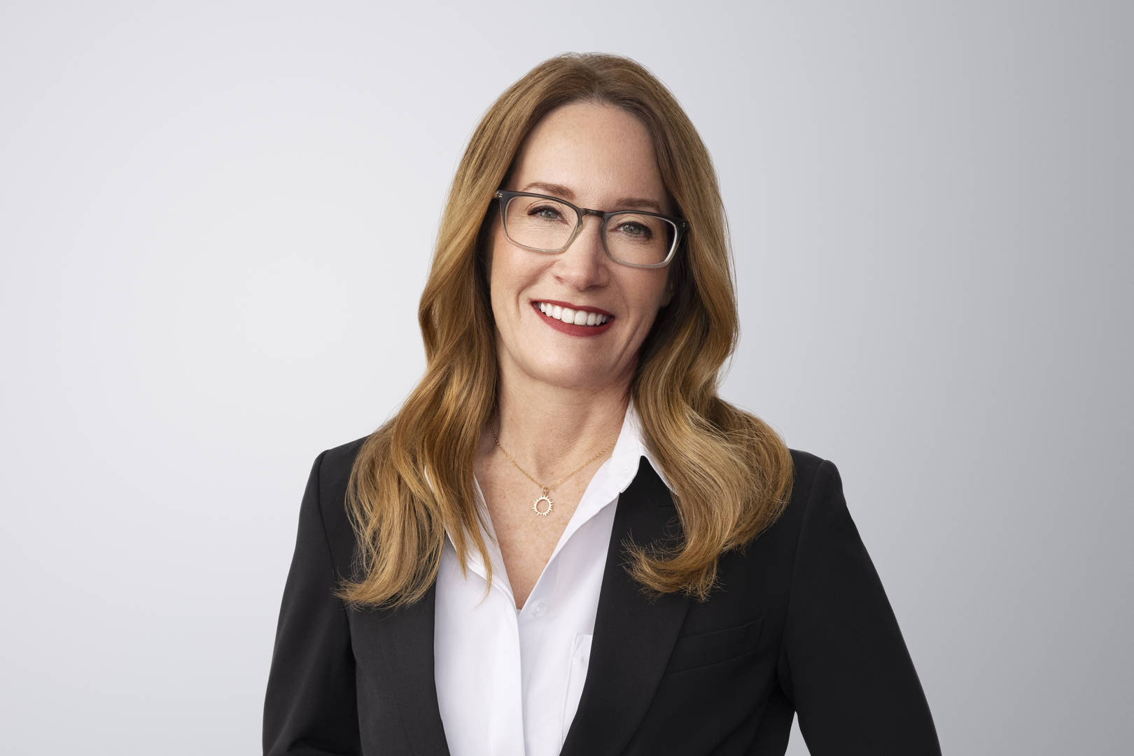 Fotografía de Elizabeth Campbell. Elizabeth, una persona de piel clara con cabello castaño y ojos verdes, sonríe a la cámara. Lleva una chaqueta blazer negra sobre una camisa blanca y un collar. Está frente a un fondo gris claro sin entornos.